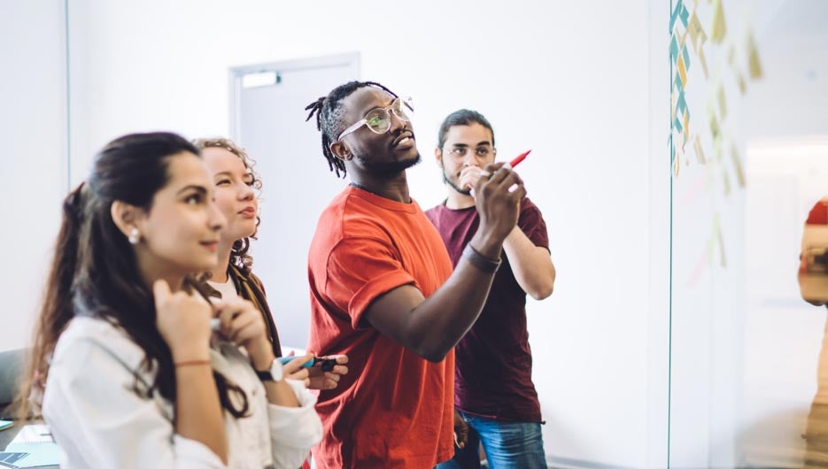 Side view of young multiracial men and women gathering in office with bright sticky notes on glass wall while planning project